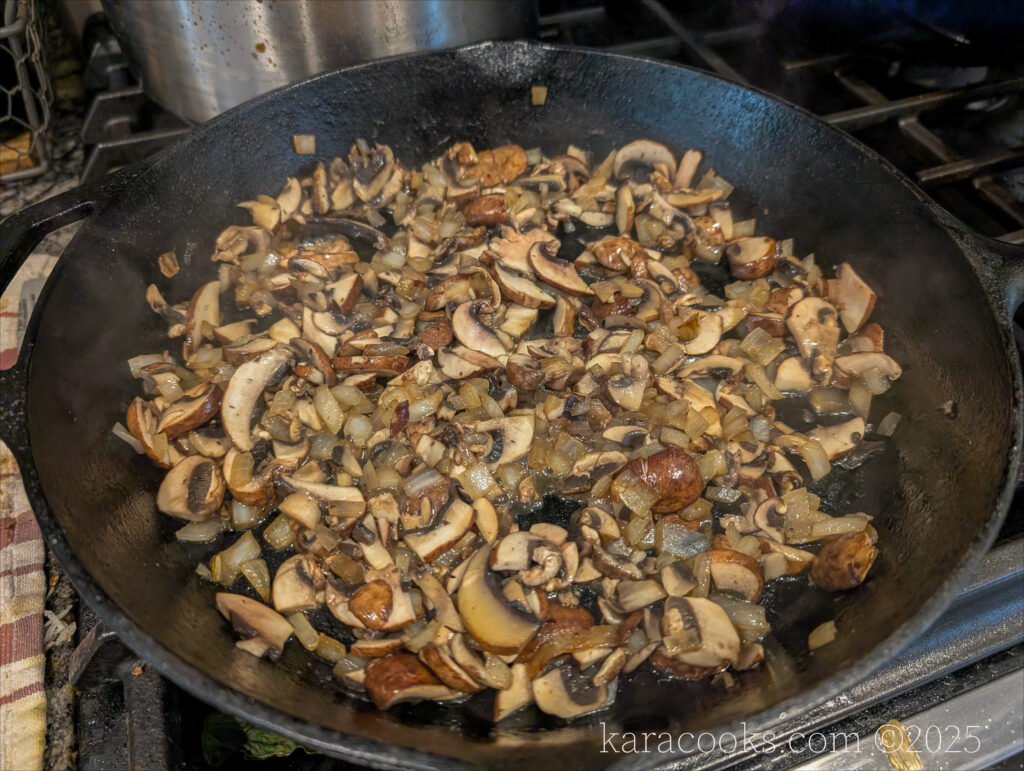 A cast iron skillet is on the stovetop filled iwth onions and mushrooms.