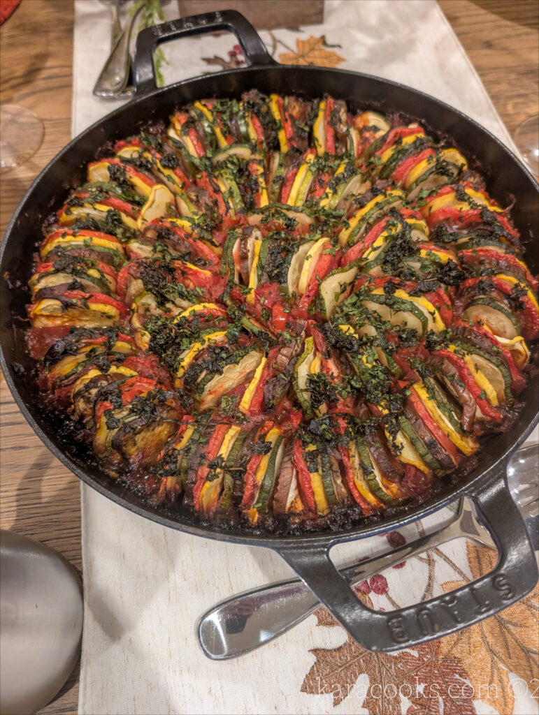 A blue Staub braiser sits on a runner embroidered with fall leaves. It's full of ratatouille and sprinkled with herbs.