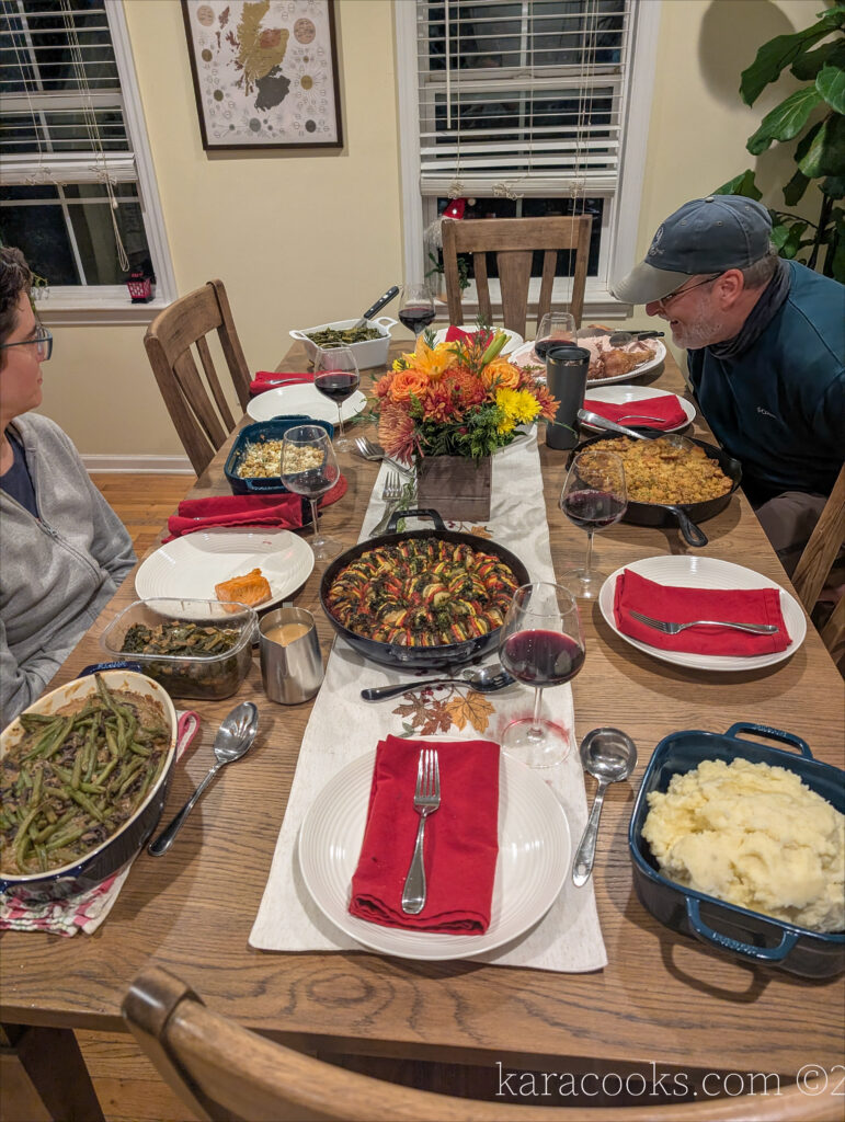 A dinner table seating 6 people is set for Thanksgiving dinner. There's a long runner embroidered with autumn leaves, a small bouquet of autumn flowers, white plates with red napkins and silverware, and a variety of dishes set around the table.