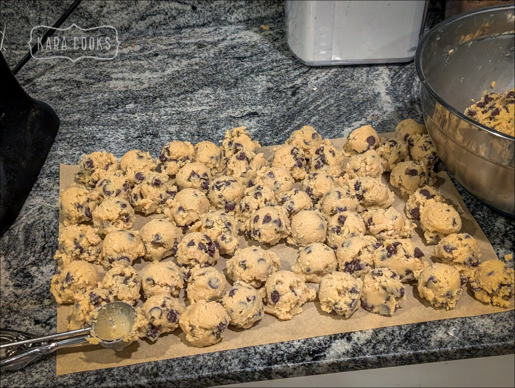 A sheet of parchment paper on a grey and white granite countertop. The parchment sheet is filled with indivudal balls of chocolate chip cookie dough. To the right of the sheet is the edge of a large bowl with more dough in it. In the front is the used cookie scoop.