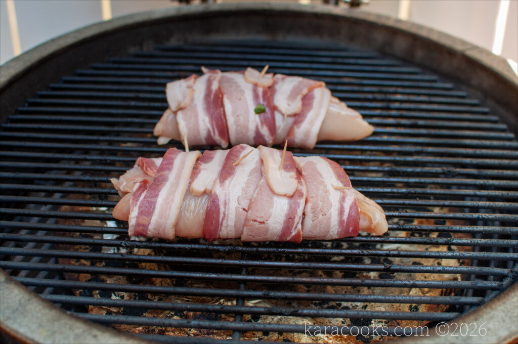 two bacon-wrapped chicken breasts on the grate of a kamado grill with glowing coals beneath