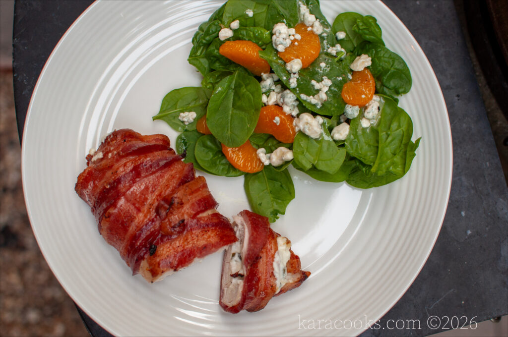 top down view of a bacon wrapped chicken breast on a plate, sliced open to show a filling of cream cheese and jalapeno bits, and a green salad with citrus.