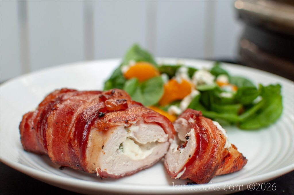 a bacon wrapped chicken breast on a plate, sliced open to show a filling of cream cheese and jalapeno bits. In the background is a green salad with citrus.