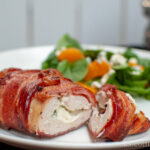a bacon wrapped chicken breast on a plate, sliced open to show a filling of cream cheese and jalapeno bits. In the background is a green salad with citrus.