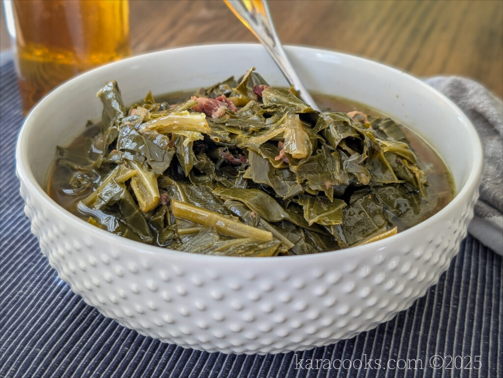 A white hobnailed bowl full of deep green braised collard greens. It's resting on a blue pin-striped placemat. There's a spoon in the bowl. There's a bottle of homemade pepper vinegar in the background.