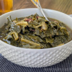 A white hobnailed bowl full of deep green braised collard greens. It's resting on a blue pin-striped placemat. There's a spoon in the bowl. There's a bottle of homemade pepper vinegar in the background.